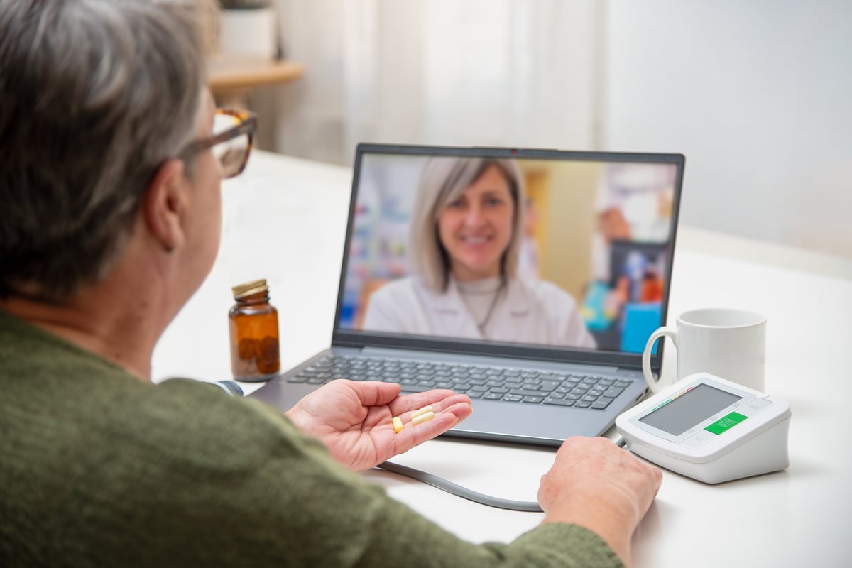 Senior adult woman showing pills to doctor during telemedicine session on laptop while measuring blood pressure